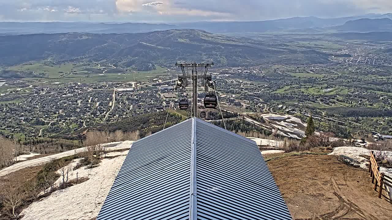 A chairlift ascends above a mountain town on a partly cloudy day, with patches of snow visible on the mountainside and a ridged metal roof in the foreground.