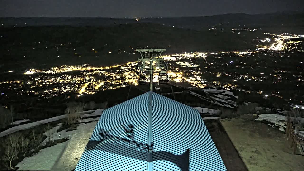 At night, a chairlift rises above a snow-dusted hillside overlooking a brightly lit town.