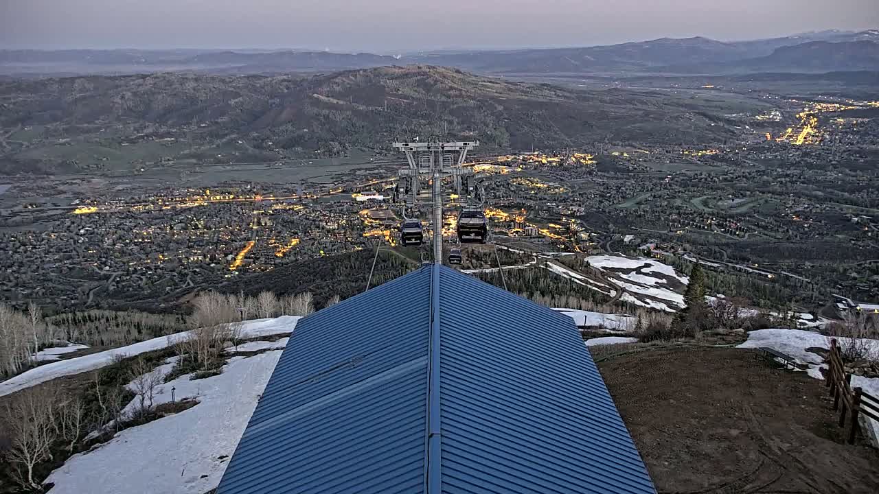 A ski lift ascends above a valley town at dusk, with patchy snow visible on the surrounding mountainside.