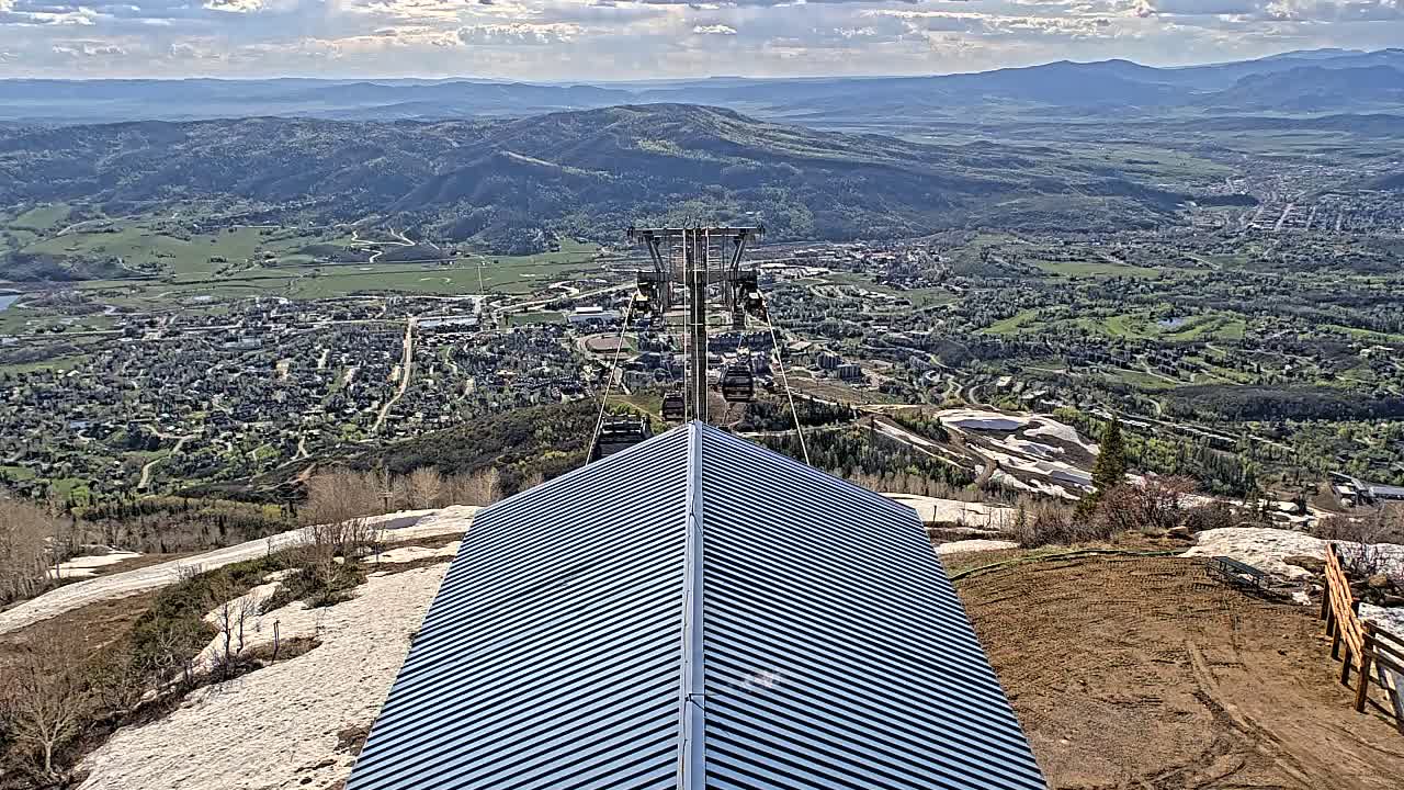 A peaked metal roof sits atop a mountain overlooking a sprawling town and valley under a partly cloudy sky.