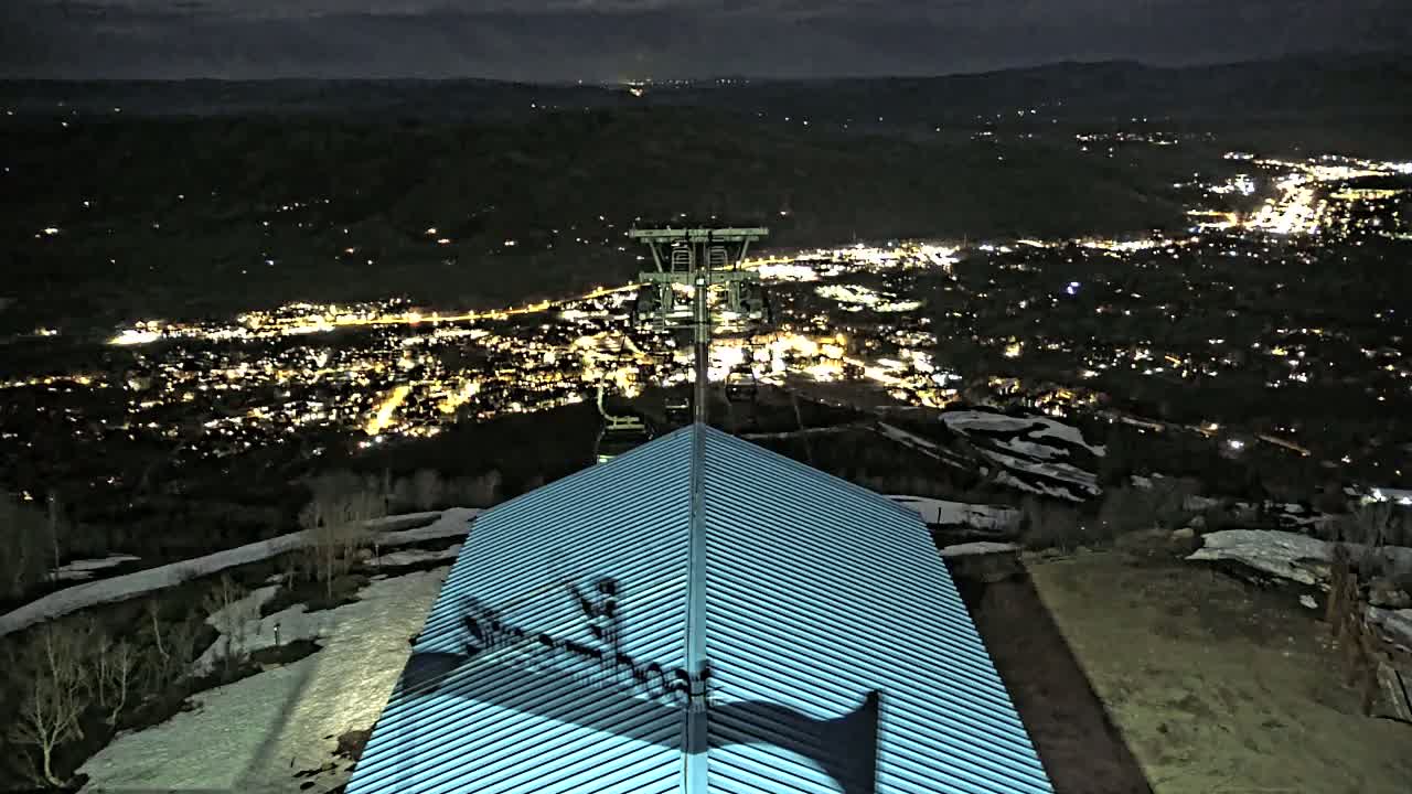 A nighttime view from a snow-dusted hilltop shows a metal roof, a ski lift, and a brightly lit town spread out below.