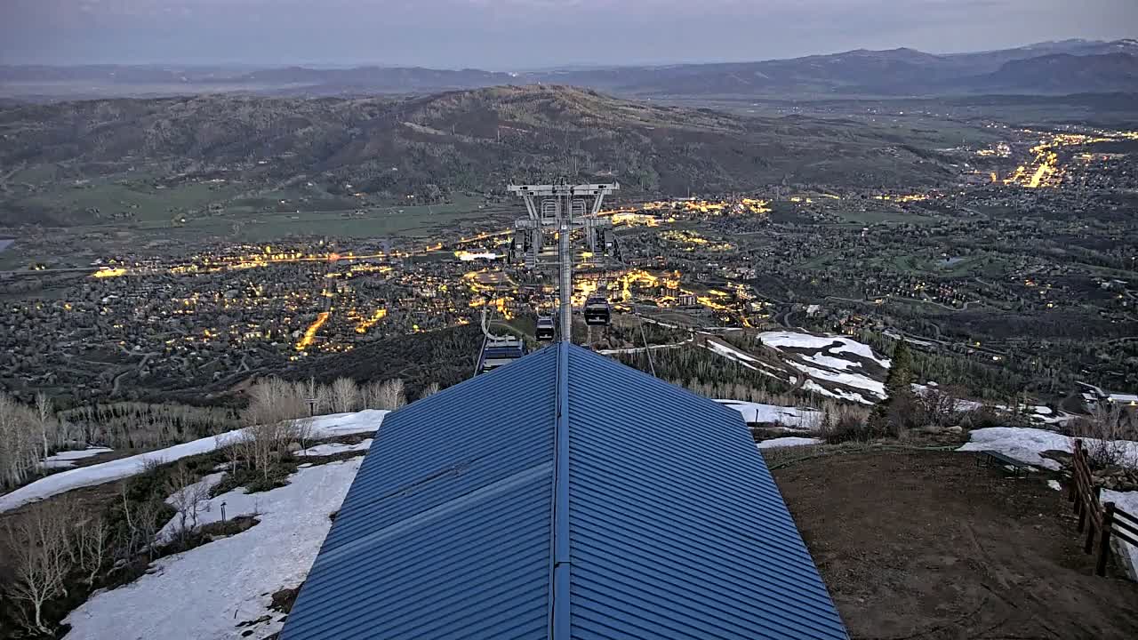 A blue roofed structure overlooks a valley town illuminated at night, with patches of snow visible on the surrounding hillside under a dusky sky.