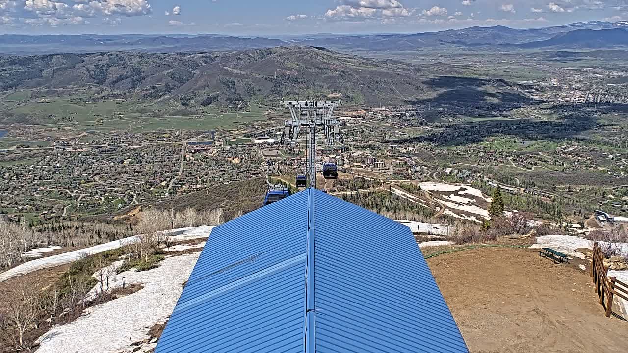 A blue-roofed building overlooks a valley town with patches of snow on the surrounding mountains under a partly cloudy sky.