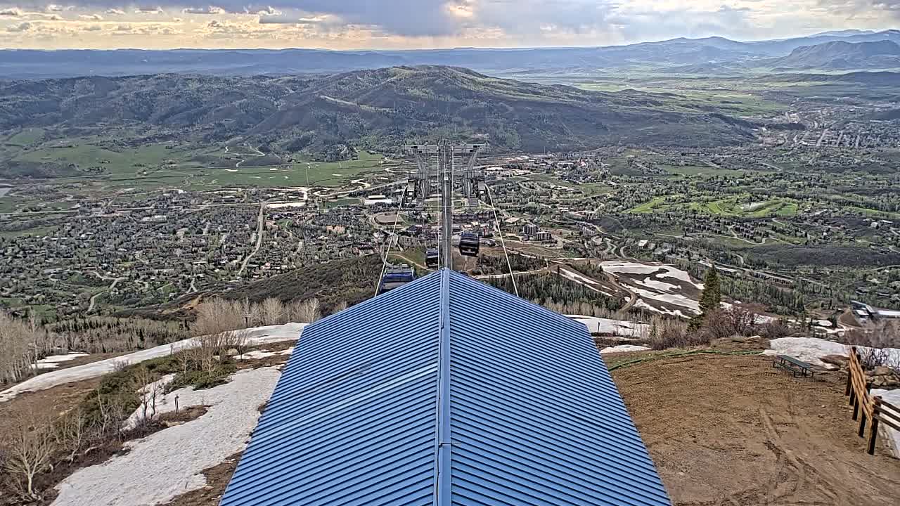 A blue roofed structure overlooks a valley town with patchy snow on the surrounding hills under partly cloudy skies.