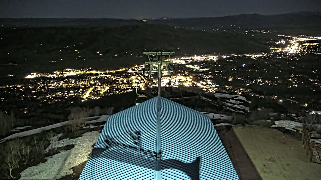 A nighttime view from a mountaintop shows a lighted town below, partially snow-covered ground nearby, and a metal roof in the foreground with a ski lift in the middle distance.