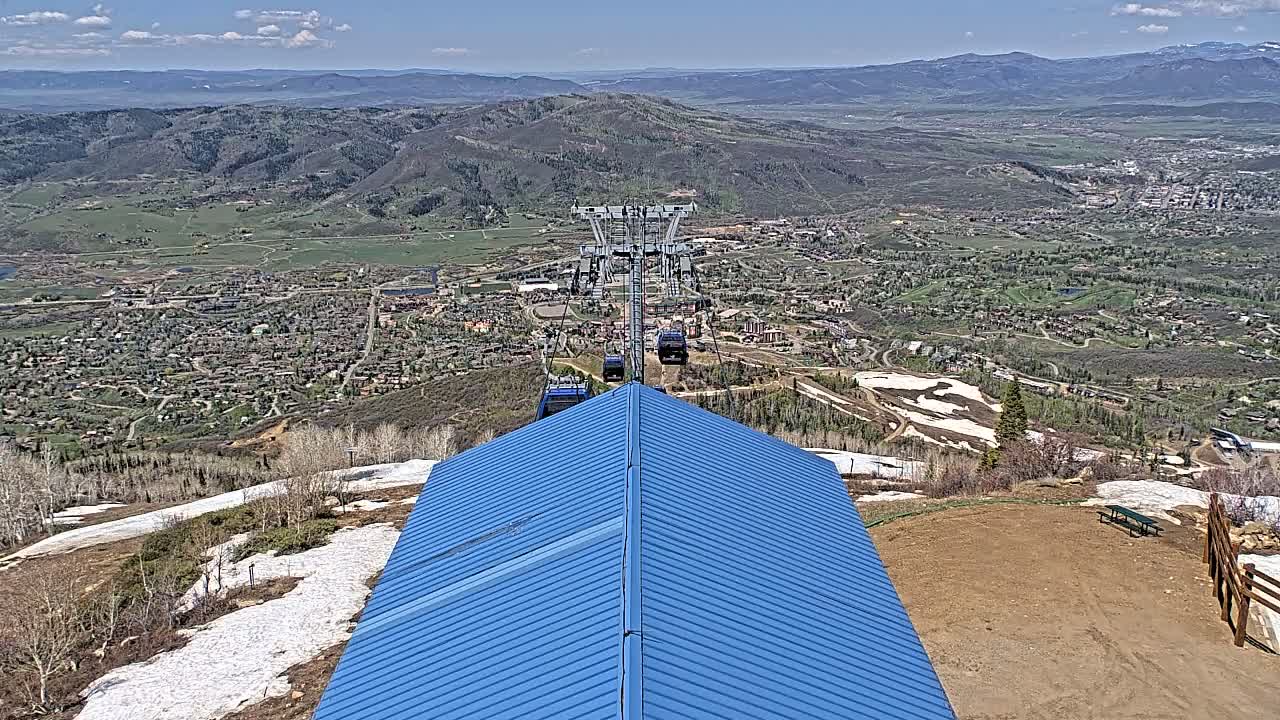 A blue metal roofed structure overlooks a valley town with patchy snow on the surrounding hills under a mostly sunny sky.