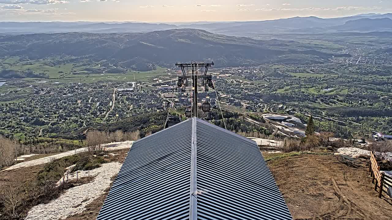 A mountaintop view reveals a chairlift ascending toward a valley town under a partly cloudy sky, with patches of snow visible on the mountainside.