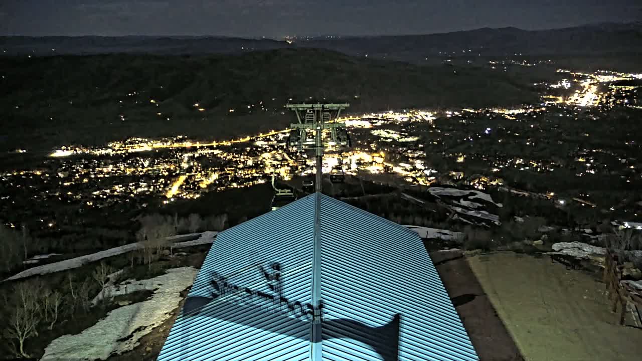 A nighttime view from a mountaintop shows a chairlift overlooking a city illuminated with lights, some patches of snow are visible on the ground, and a light-blue metal roof is in the foreground.