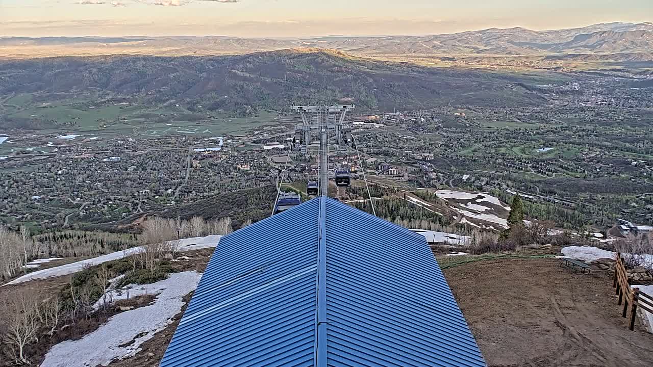 A blue-roofed structure sits atop a mountain overlooking a valley town with patches of snow on the ground, under a mostly clear sky.