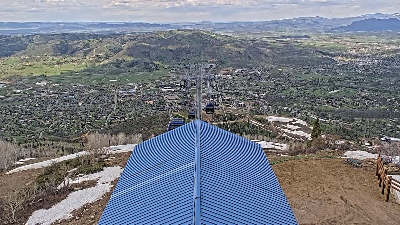 A blue-roofed building overlooks a valley town with a chairlift ascending into the partly cloudy sky, with patches of snow still visible on the mountainside.