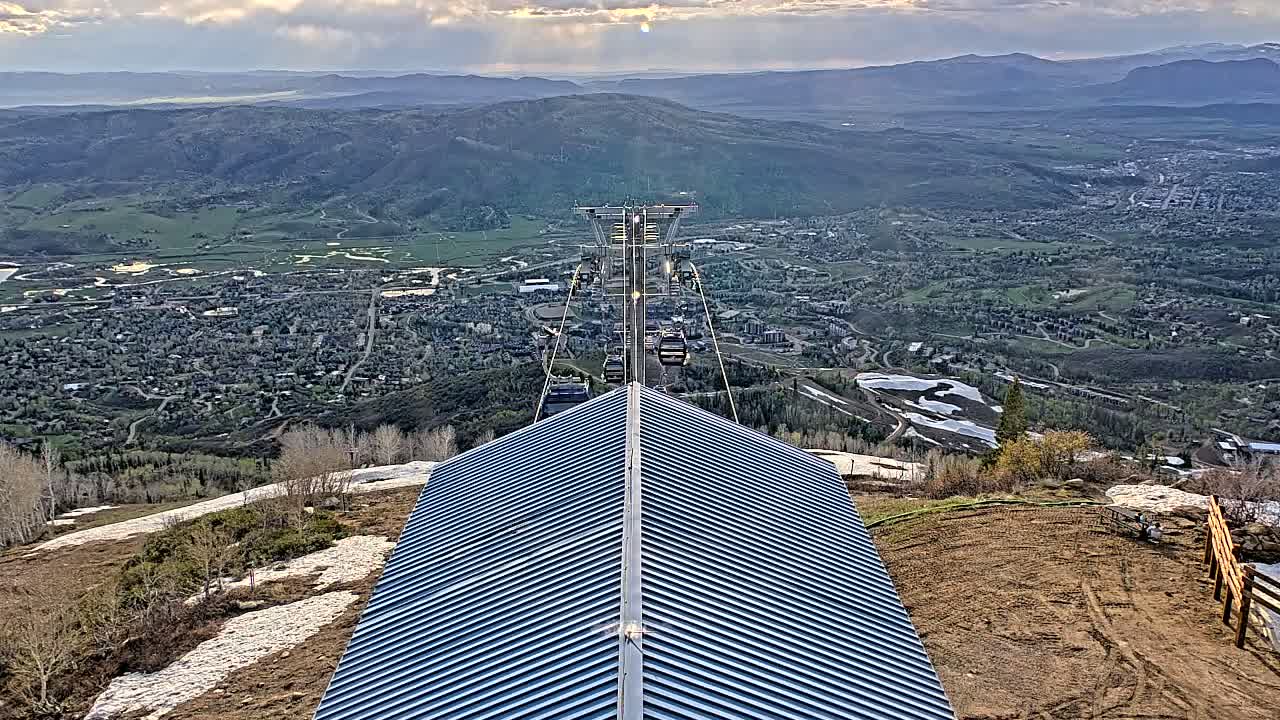 A mountaintop view shows a chairlift descending toward a town nestled in a valley, with patches of snow visible on the mountainside under partly cloudy skies.