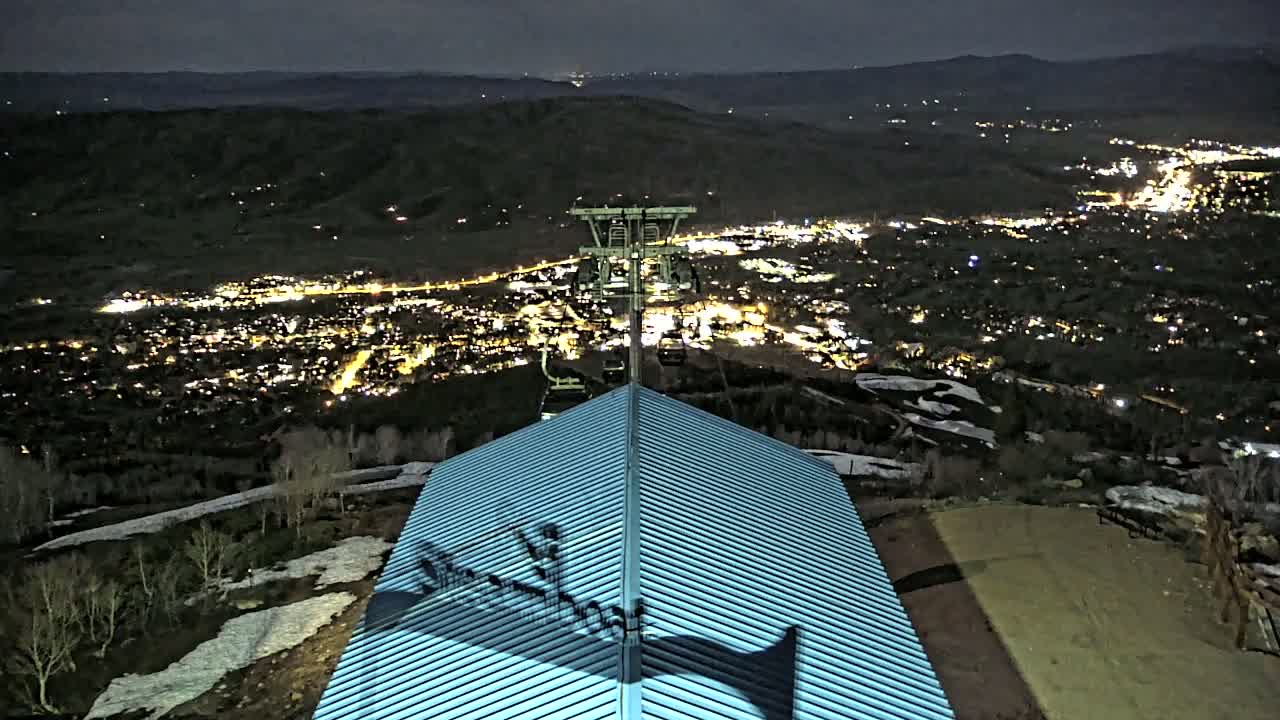 A nighttime view from a mountaintop shows a chairlift overlooking a city illuminated by lights, with patches of snow visible on the ground and a peaked, light-blue roof in the foreground.