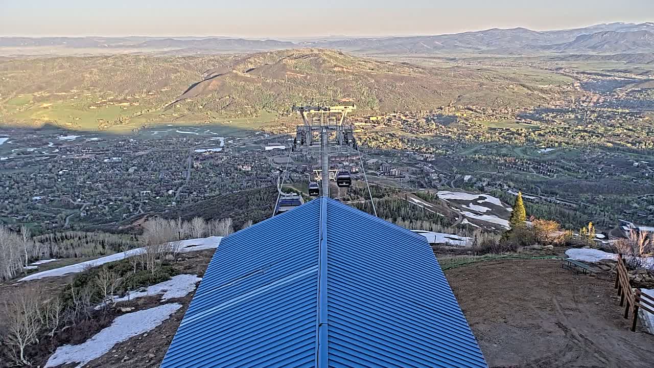 A blue metal roofed structure overlooks a mountain valley town with patches of snow on the surrounding hillsides under a clear sky.