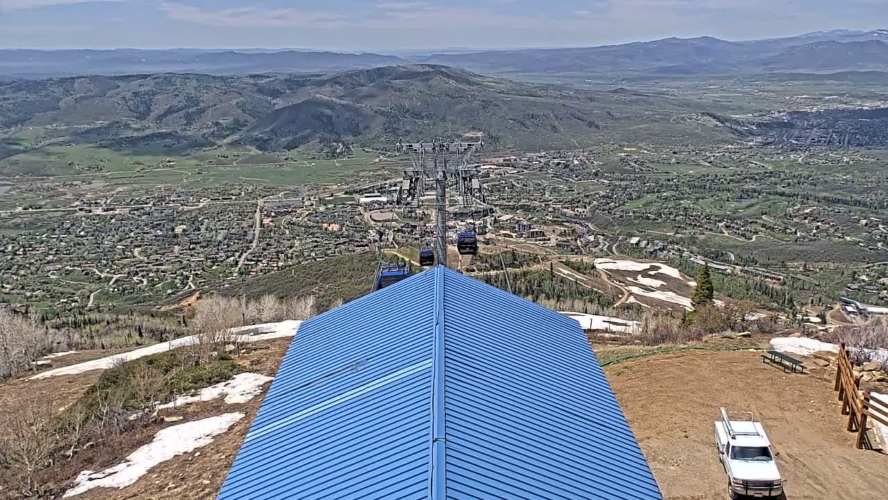 A blue-roofed structure sits atop a mountain overlooking a valley town under a clear, sunny sky, with a chairlift carrying passengers visible in the distance and patches of remaining snow on the mountainside.