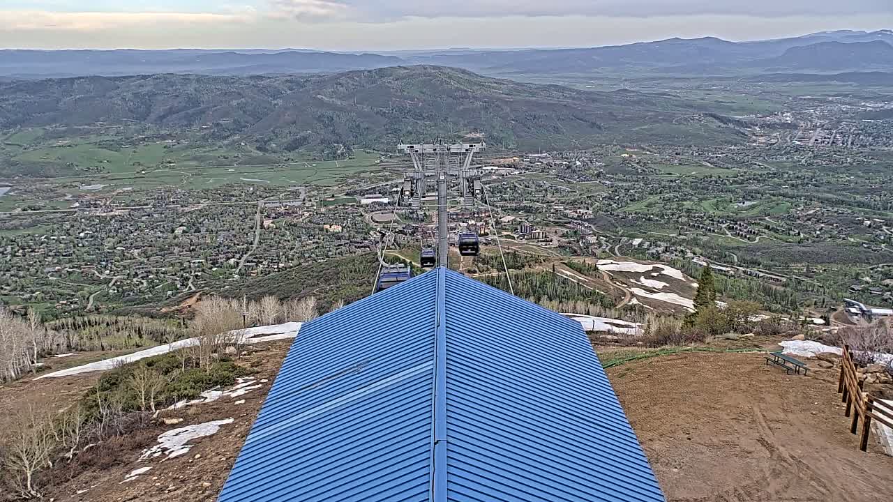 A blue roof is situated on a mountaintop overlooking a valley town, with a chairlift ascending into the distance, under partly cloudy skies and patchy snow on the ground.