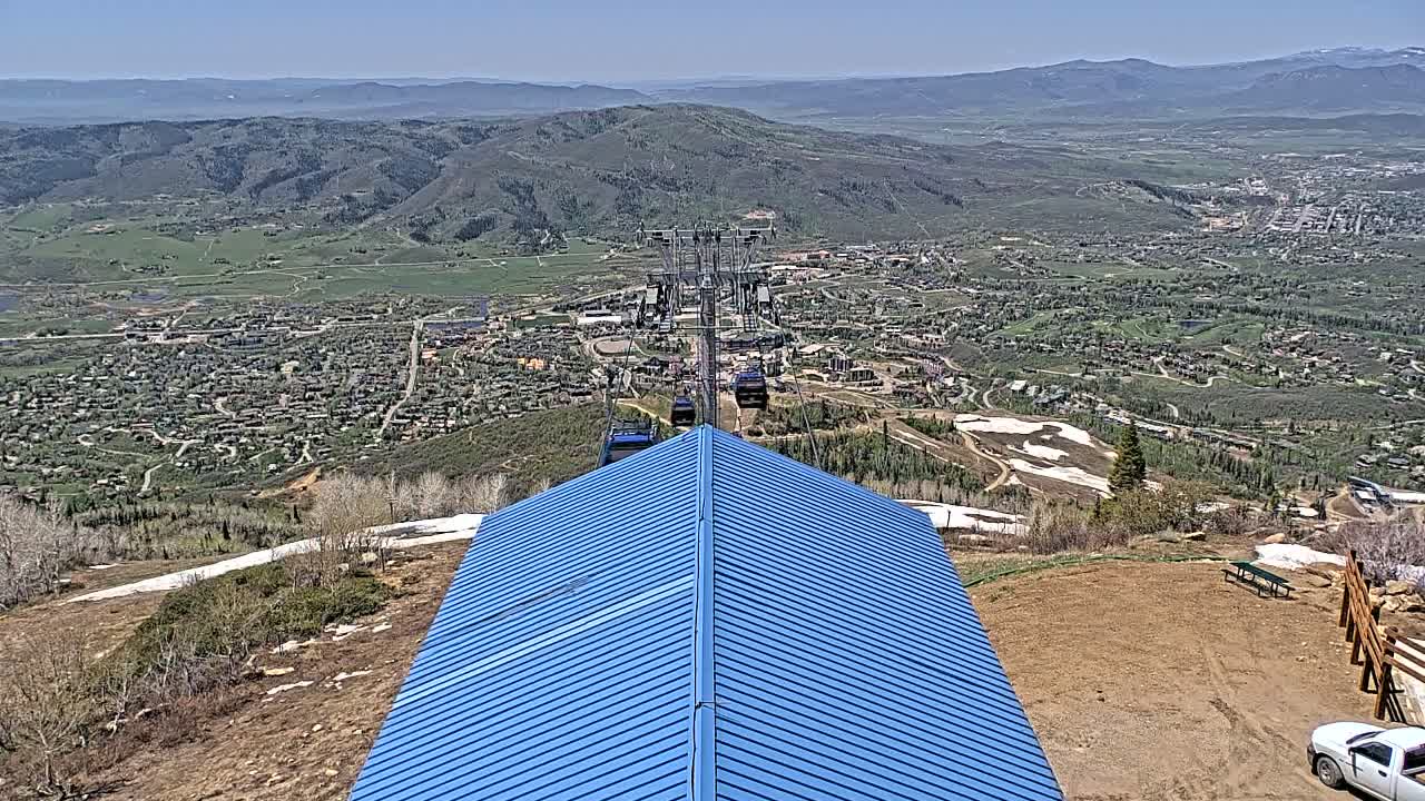 A blue-roofed structure sits atop a mountain overlooking a valley town on a clear, sunny day, with remnants of snow visible on the mountainside and in the distance a chairlift carrying passengers.