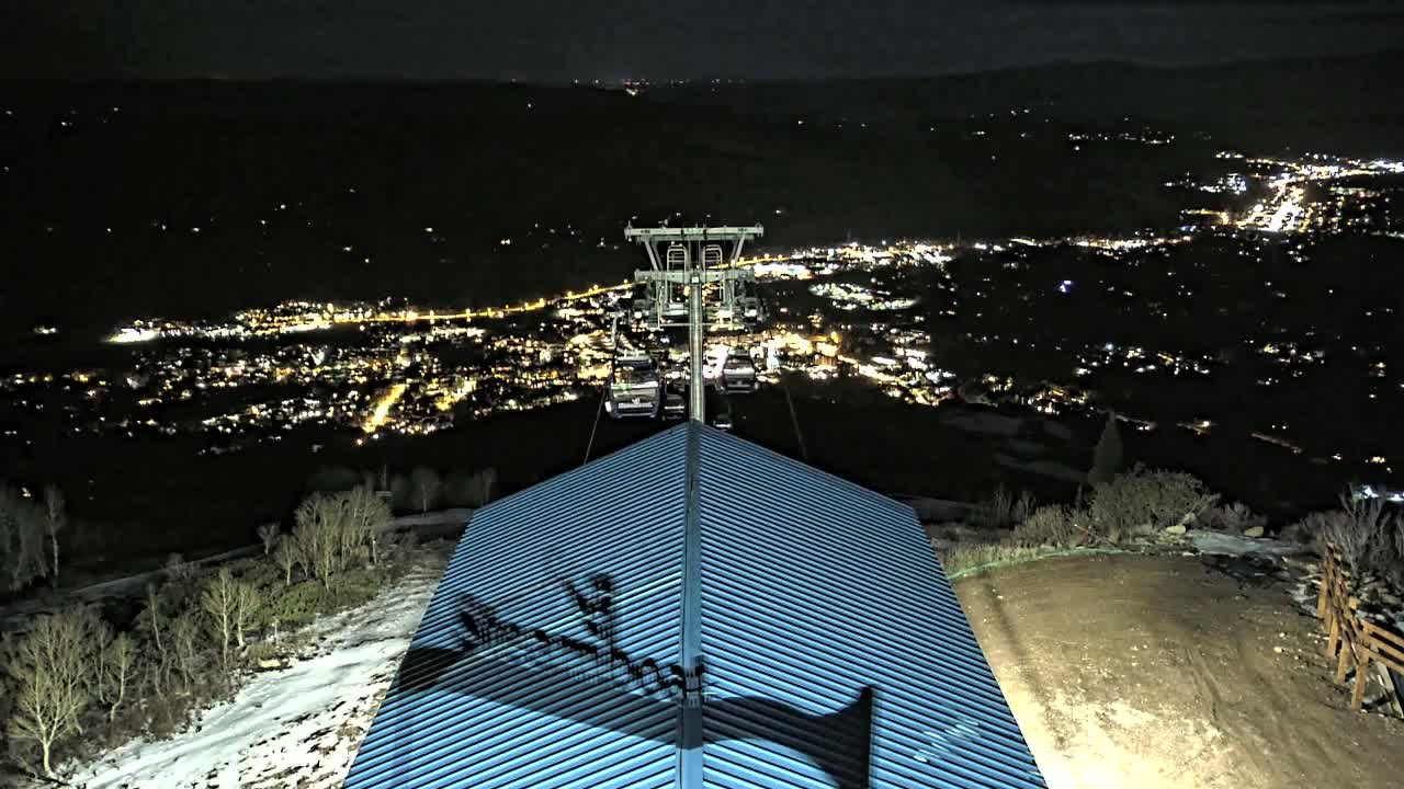 A chairlift ascends from a building with a ribbed blue roof towards a nighttime city twinkling with lights, with some snow visible on the ground.