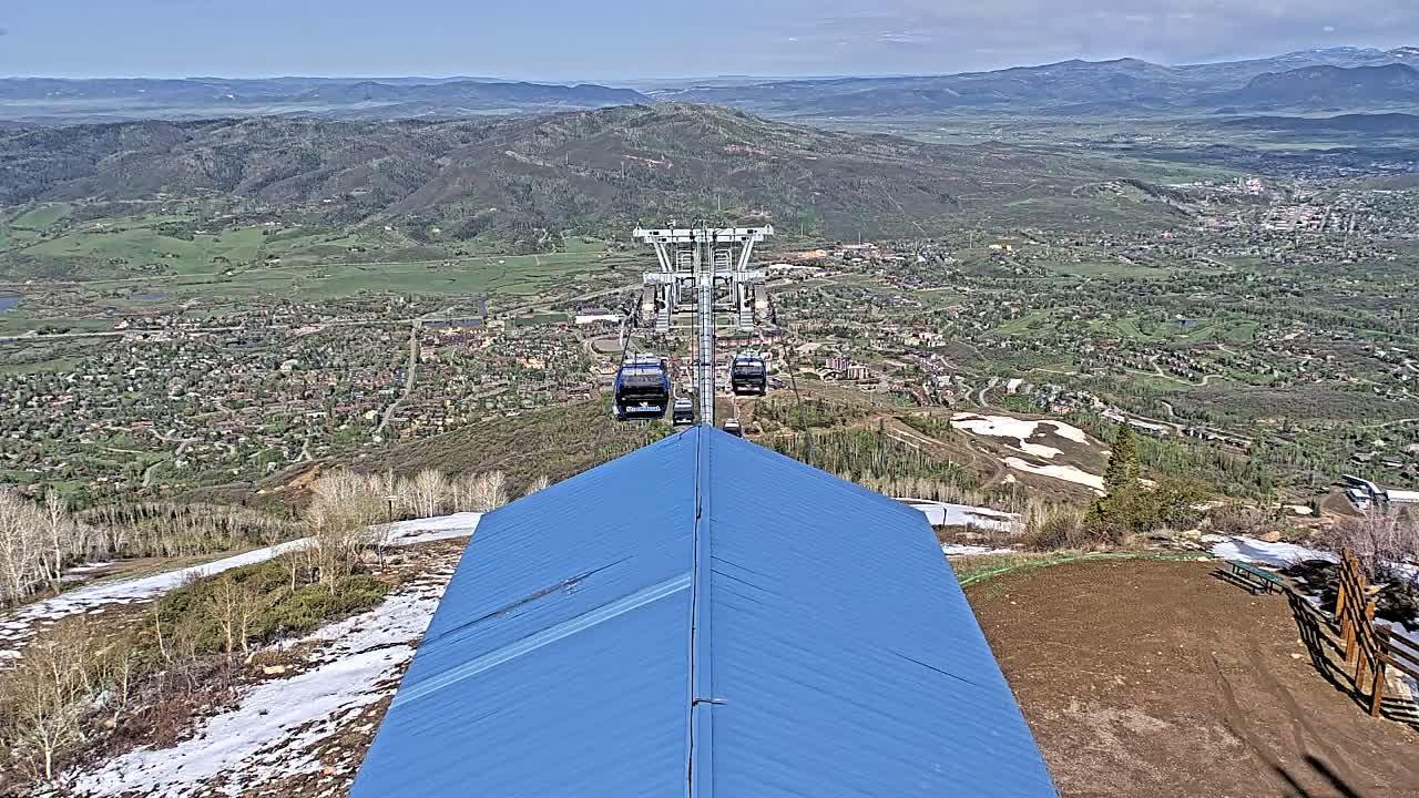 A chairlift ascends a mountain overlooking a valley town, with patches of snow visible on the mountainside under a clear sky.