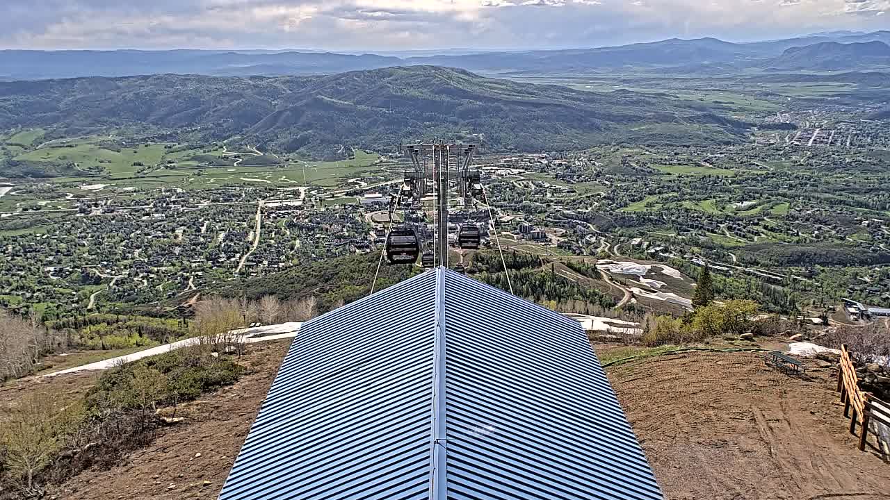 A blue metal roofed structure sits atop a mountain overlooking a valley town under partly cloudy skies, with a chairlift ascending from the valley below.