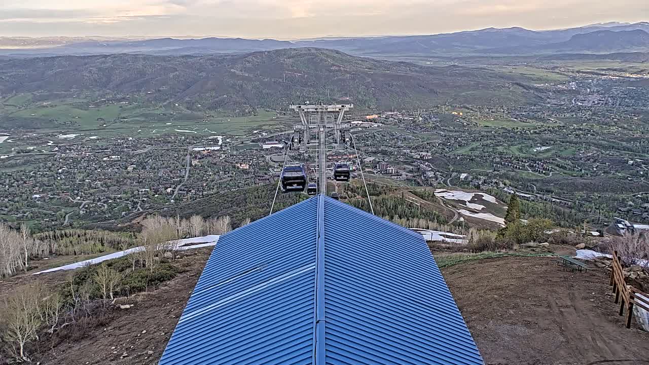 A blue-roofed structure sits atop a mountain overlooking a valley town, with a chairlift carrying several gondolas in the distance under a partly cloudy sky.