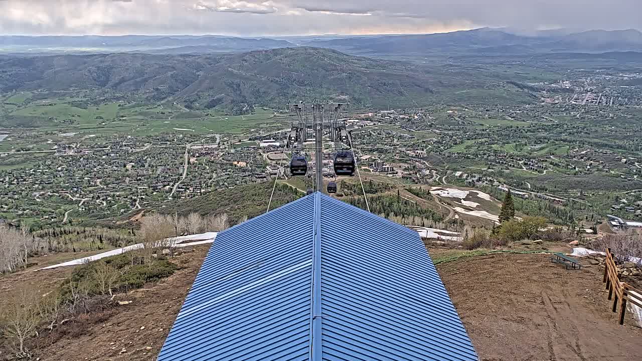 A blue-roofed structure overlooks a valley town under a partly cloudy sky, with a chairlift carrying several gondolas in the distance.