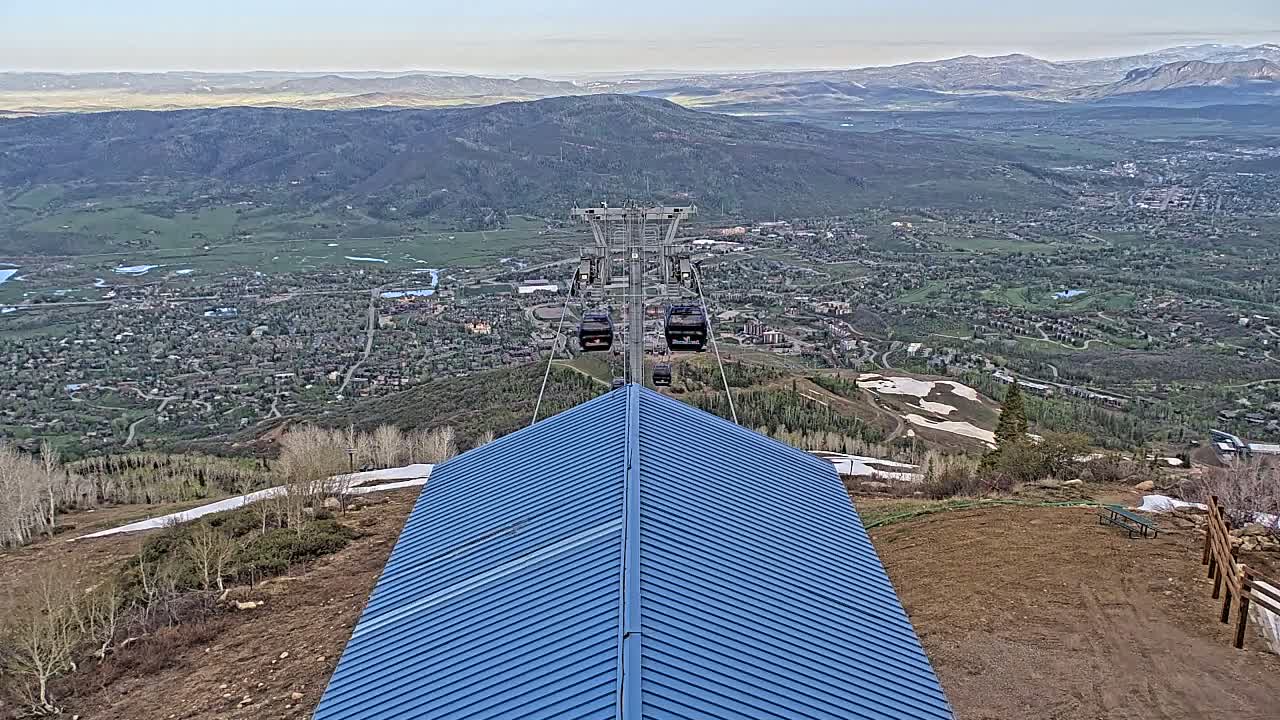 A blue-roofed structure sits atop a mountain overlooking a valley town on a clear day, with a chairlift visible in the distance.