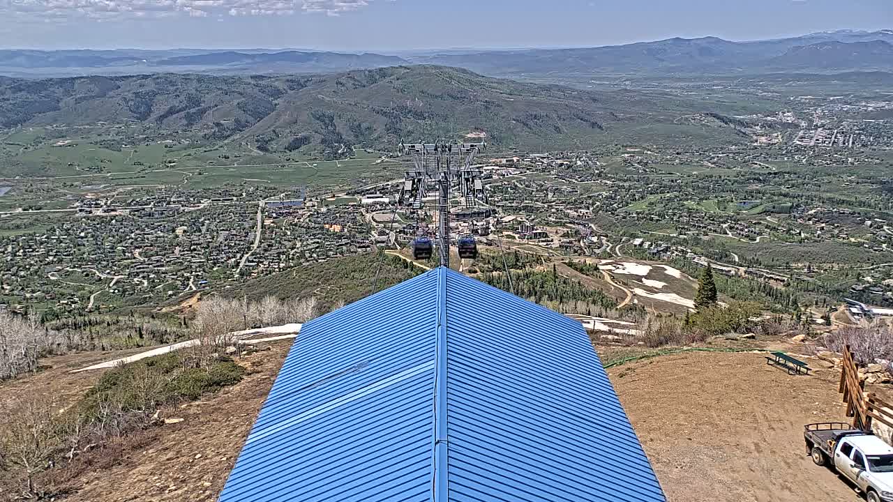 A blue-roofed structure overlooks a valley town with a chairlift ascending from the foreground on a partly cloudy day.