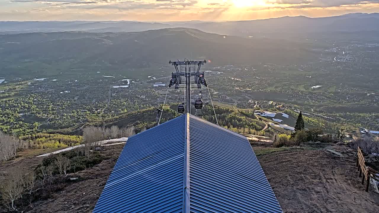 A chairlift ascends a mountain at sunset, overlooking a valley town with patches of snow visible on the slopes.