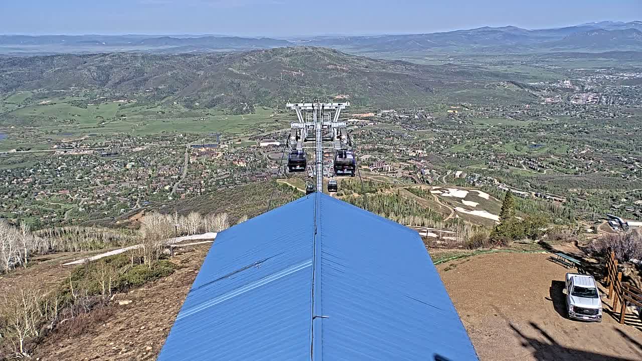 A mountaintop view on a clear day shows a chairlift descending towards a town nestled in a valley, with a blue roof and a pickup truck visible in the foreground.