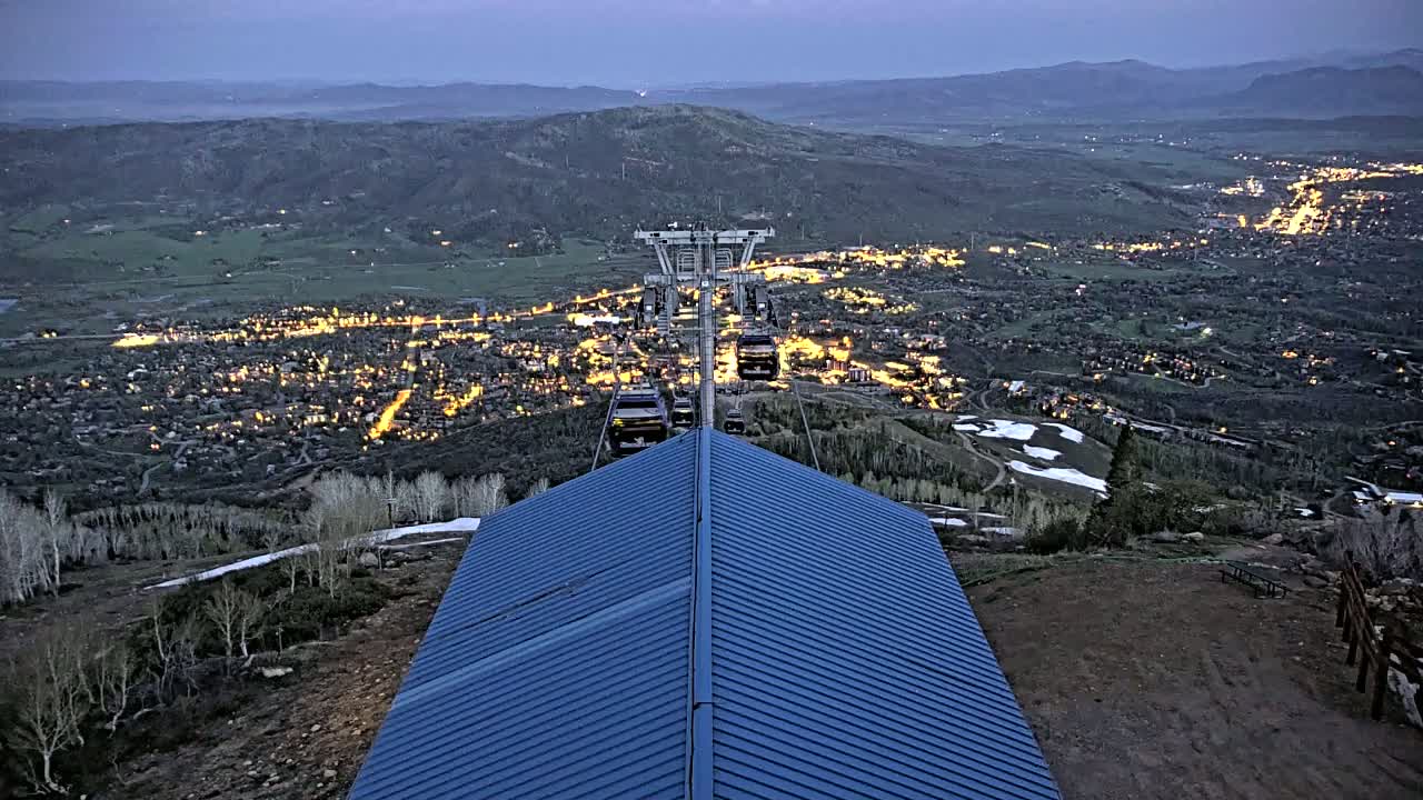 A chairlift ascends a mountain overlooking a valley town illuminated at dusk, with patches of snow visible on the mountainside.