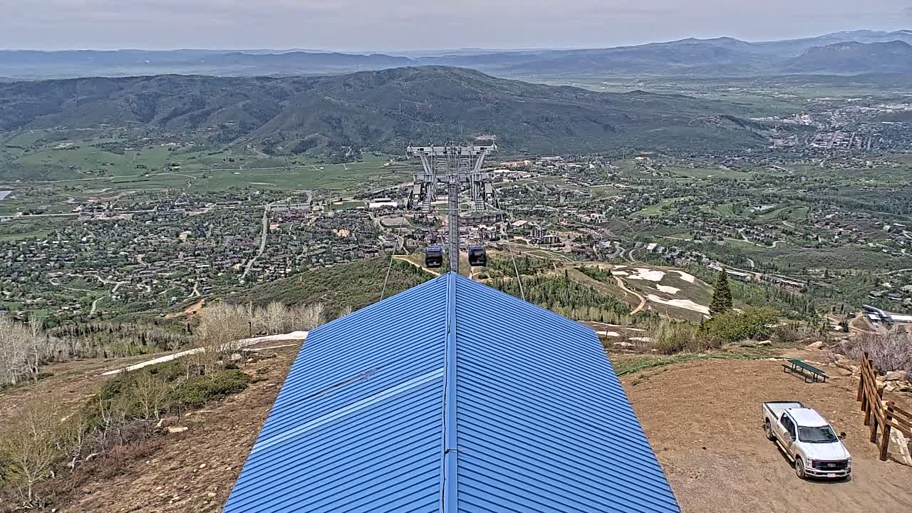 A blue peaked roof sits atop a mountain overlooking a valley town under a partly cloudy sky, with a chairlift in the distance and a truck parked nearby.