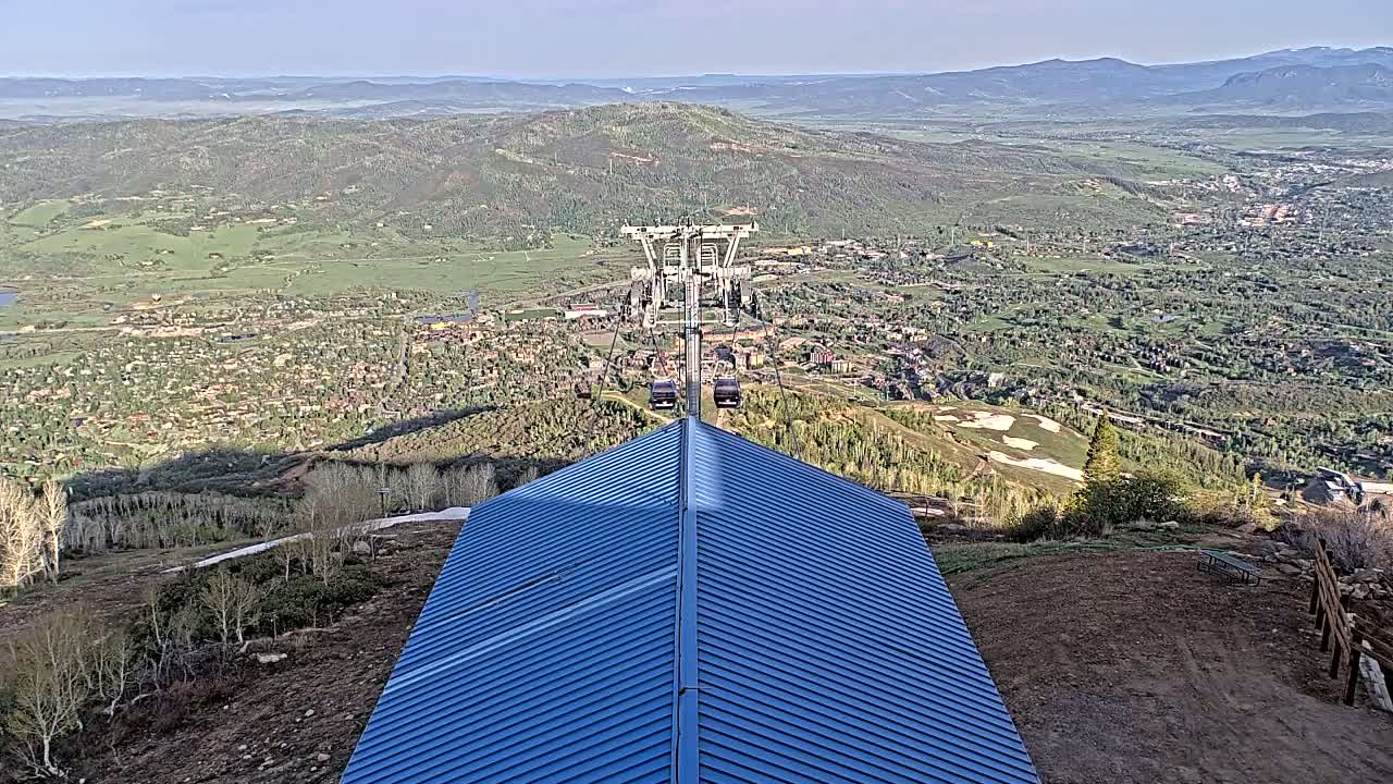A blue metal roofed structure sits atop a hill overlooking a valley town under a clear sky.