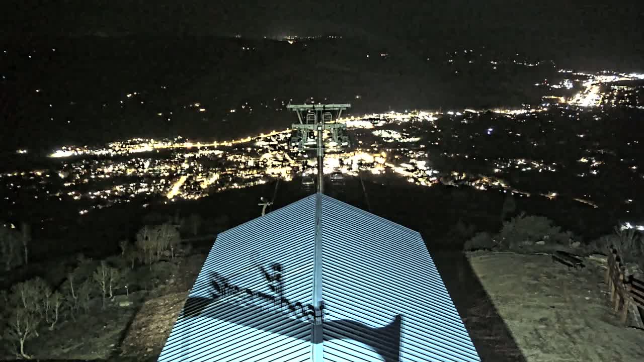 A nighttime view from a hilltop shows a metal roof with shadows cast upon it, a ski lift tower in the distance, and a brightly lit town spread out below.