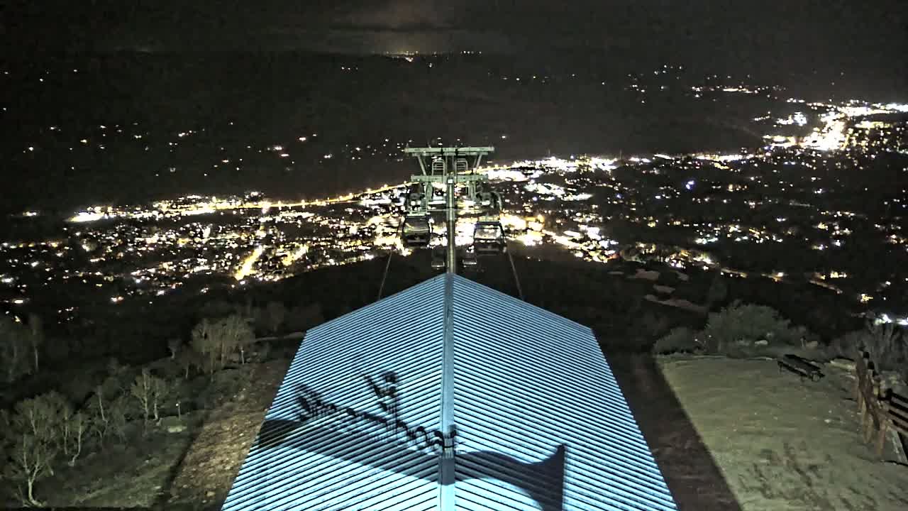 A nighttime view from a hilltop shows a chairlift ascending toward a brightly lit city in the distance, with its shadow cast on a building's roof below.