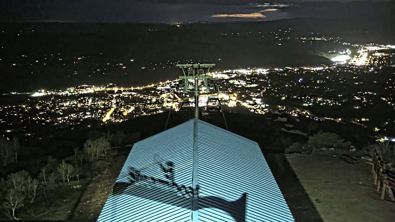A nighttime view from a mountaintop shows a city illuminated below, with a chairlift and a building's roof in the foreground.