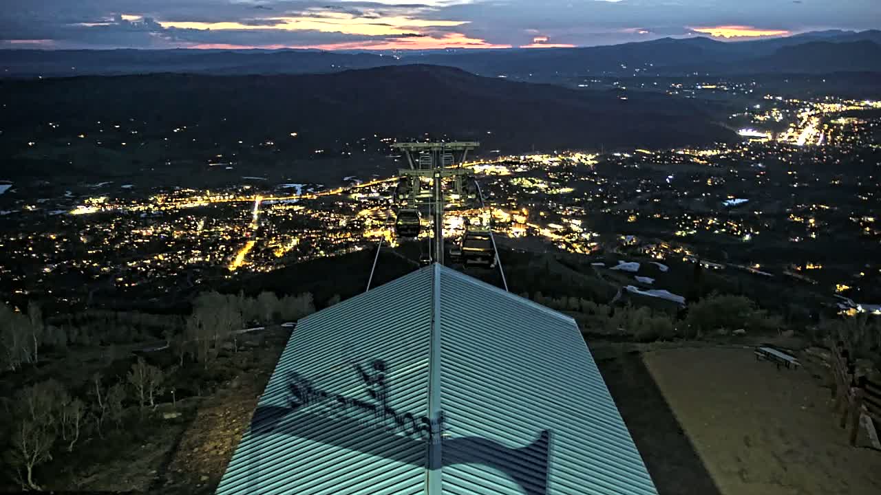 A nighttime view from a mountaintop shows a chairlift overlooking a valley town illuminated by lights, with a metallic roof in the foreground under a dusky sky.