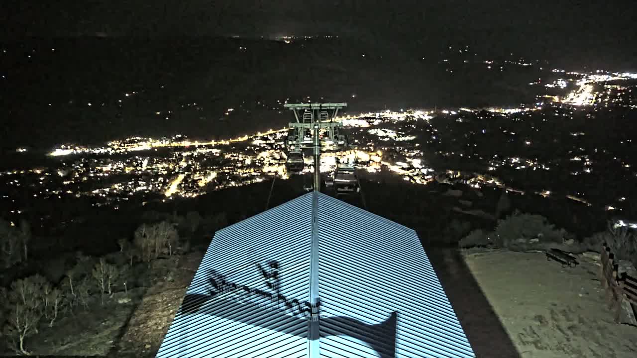 A nighttime view from a mountaintop shows a chairlift ascending toward a brightly lit city below, with a building's roof in the foreground.