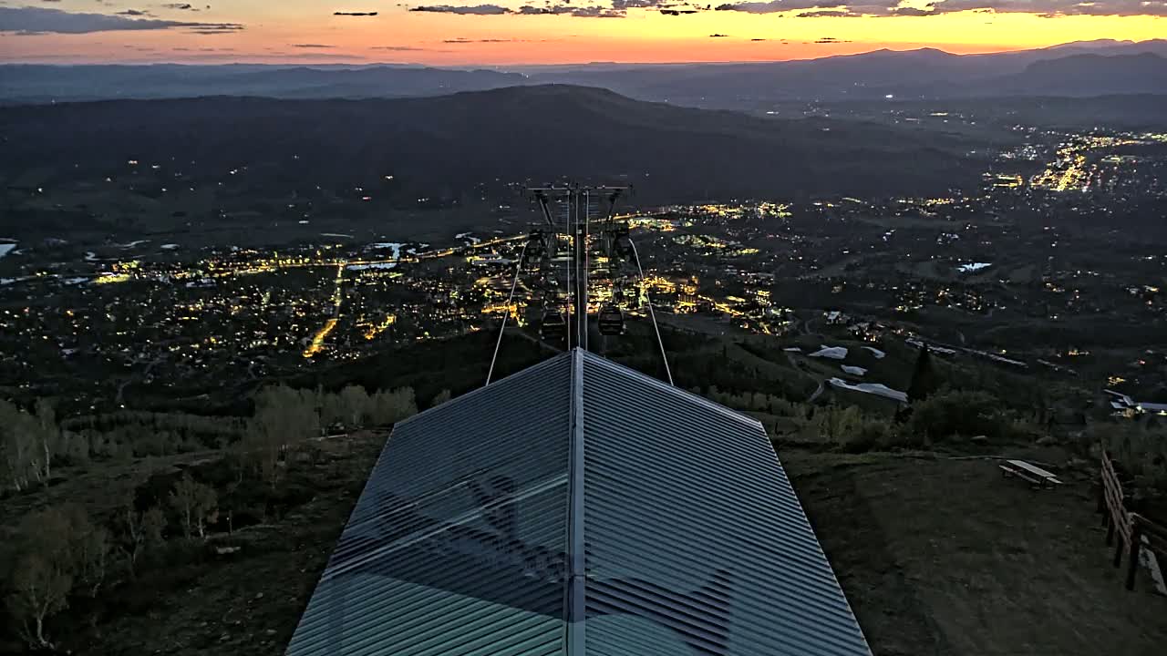 A nighttime view from a mountaintop shows a city illuminated below, with a chairlift extending down to a sloped, metallic structure, under a sunset sky.