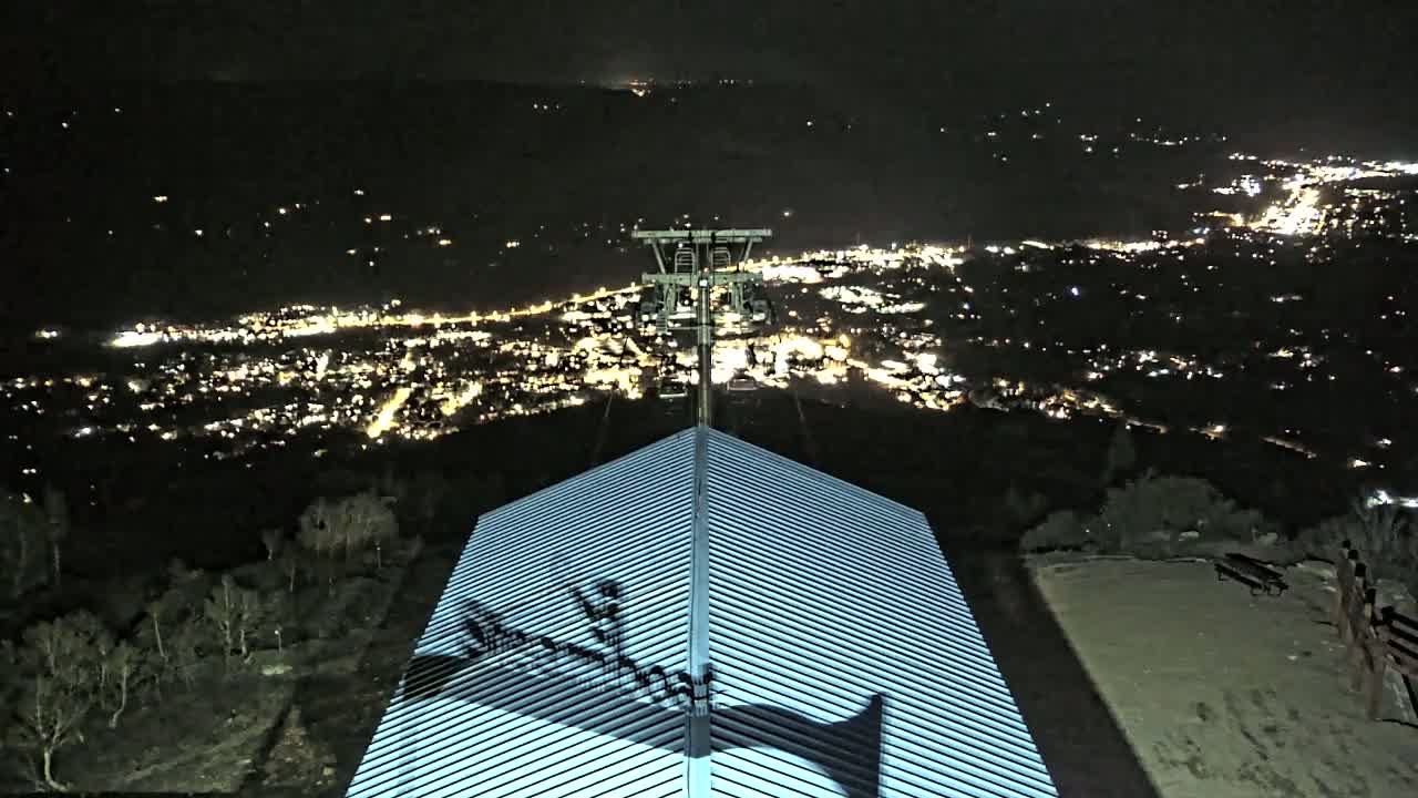 At night, a chairlift stands atop a ridged roof overlooking a brightly lit city nestled in a valley.
