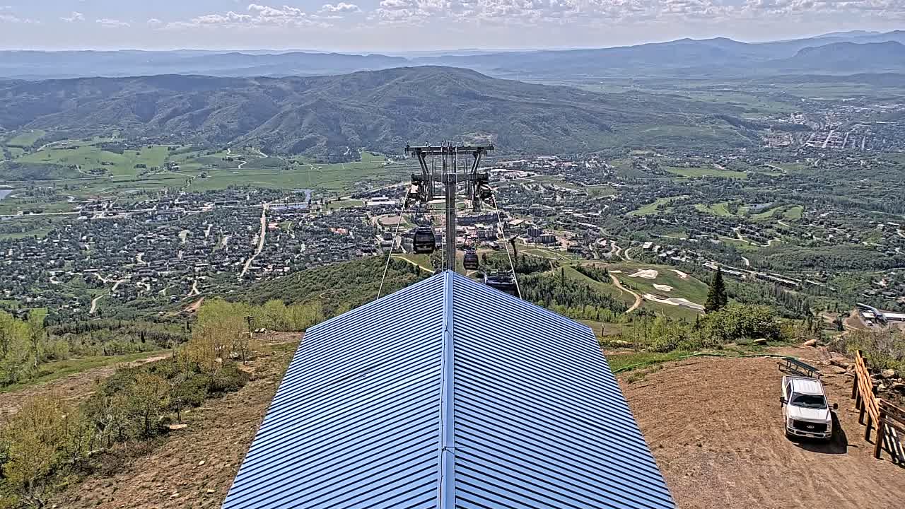 A blue metal roof of a building overlooks a valley town and distant mountains under a mostly sunny sky, with a chairlift visible in the background.