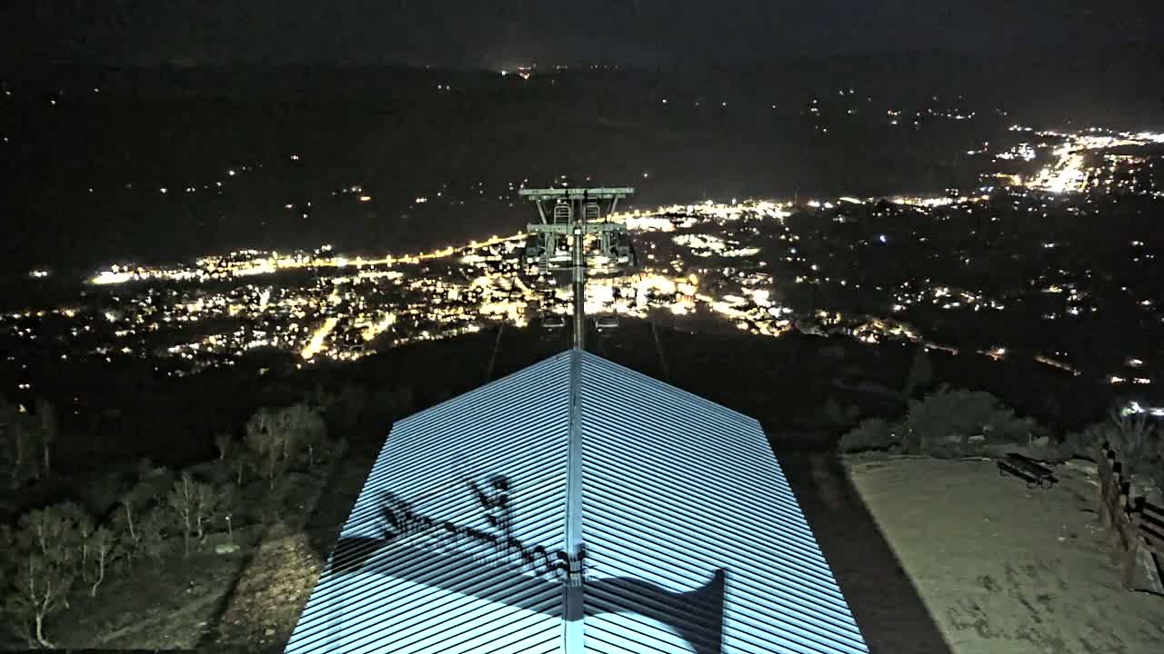 A nighttime view from a hilltop shows a chairlift ascending towards a distant city illuminated by many lights.