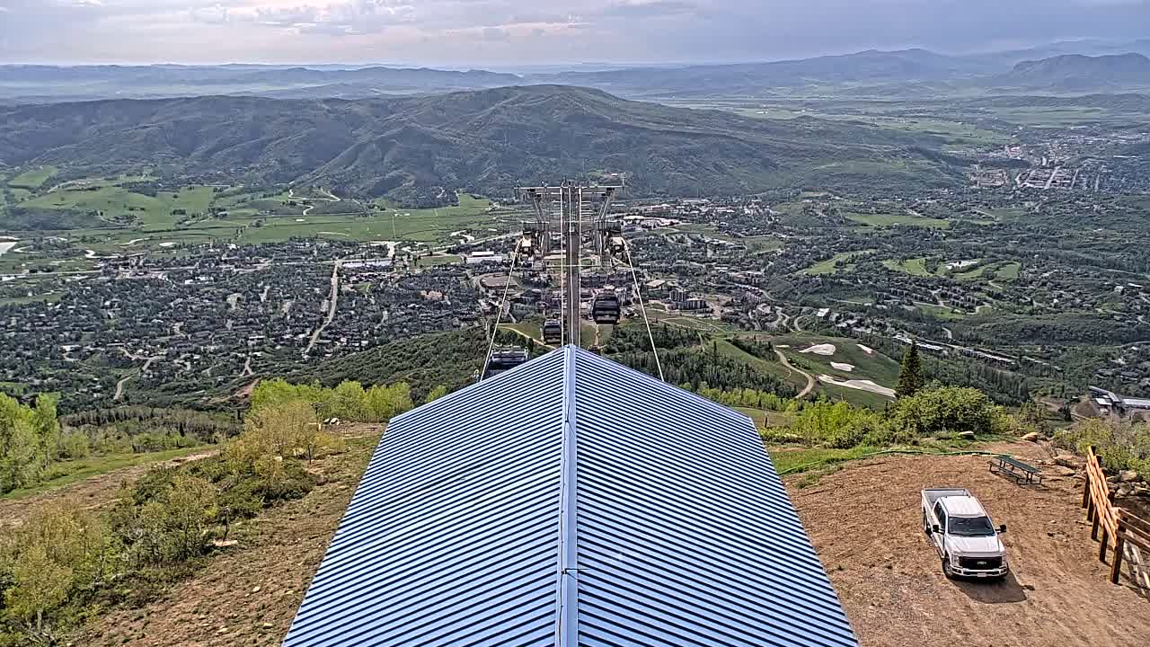 A blue-roofed structure sits atop a mountain overlooking a valley town under partly cloudy skies, with a chairlift ascending into view.