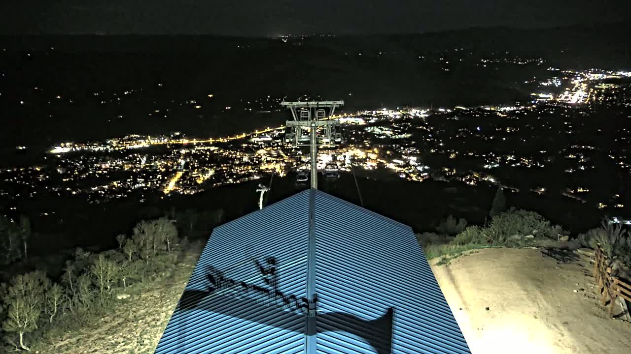 A nighttime view from atop a building shows a chairlift ascending towards a distant, brightly lit city sprawling across a valley.