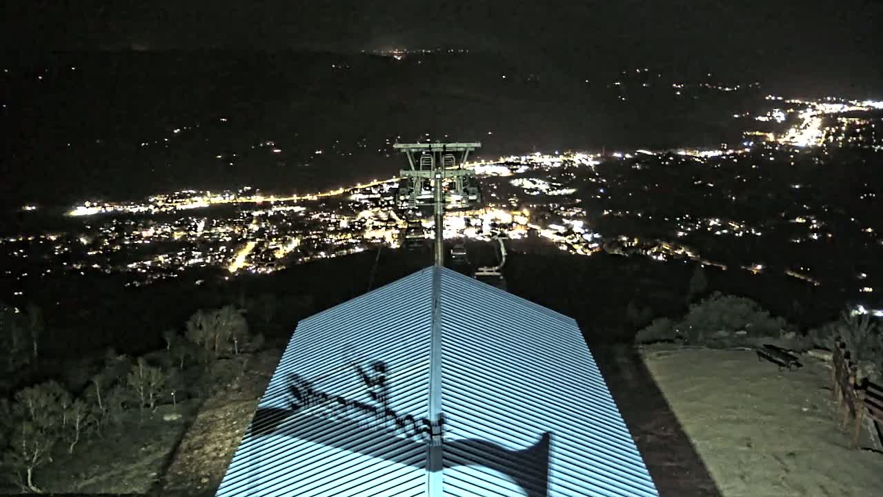 A nighttime view from a hilltop shows a city illuminated below, with a chairlift and a building's corrugated metal roof in the foreground.