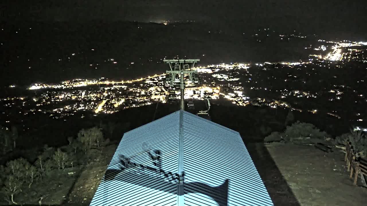At night, a building's ridged roof is illuminated, overlooking a valley filled with the lights of a distant town and a chairlift structure.