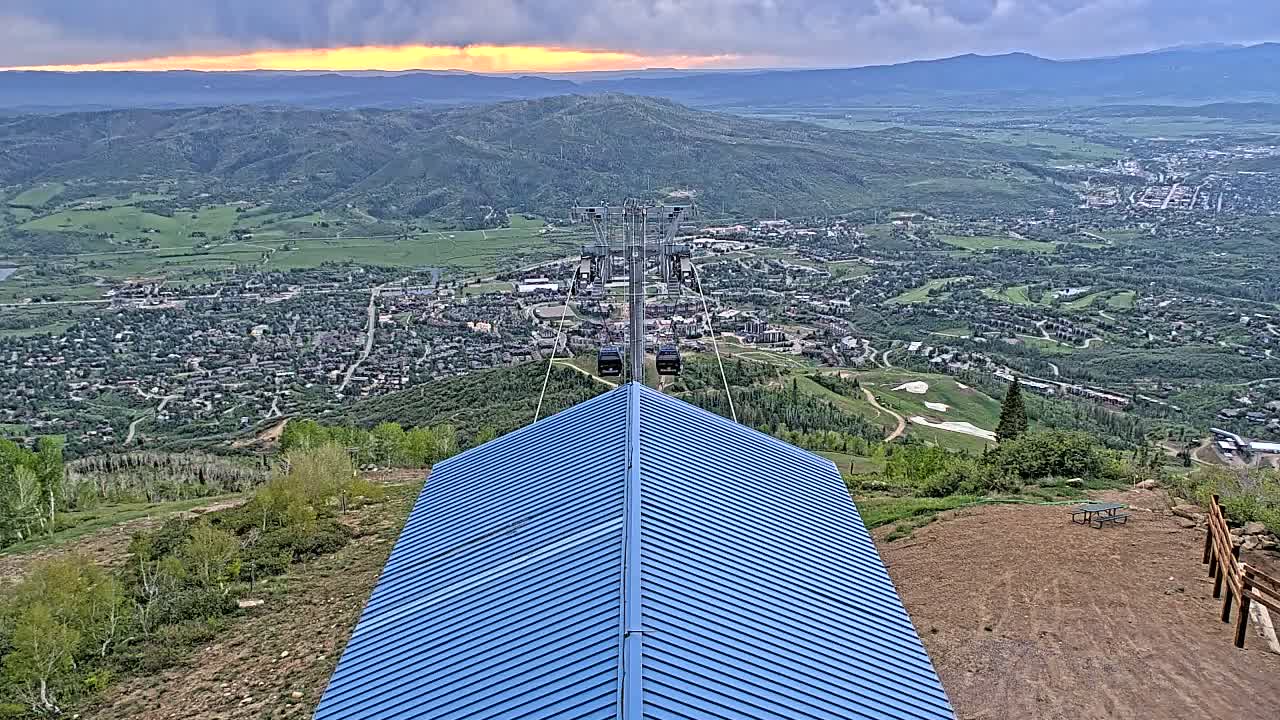 A blue metal roof sits atop a hill overlooking a valley town under a partly cloudy sunset sky.