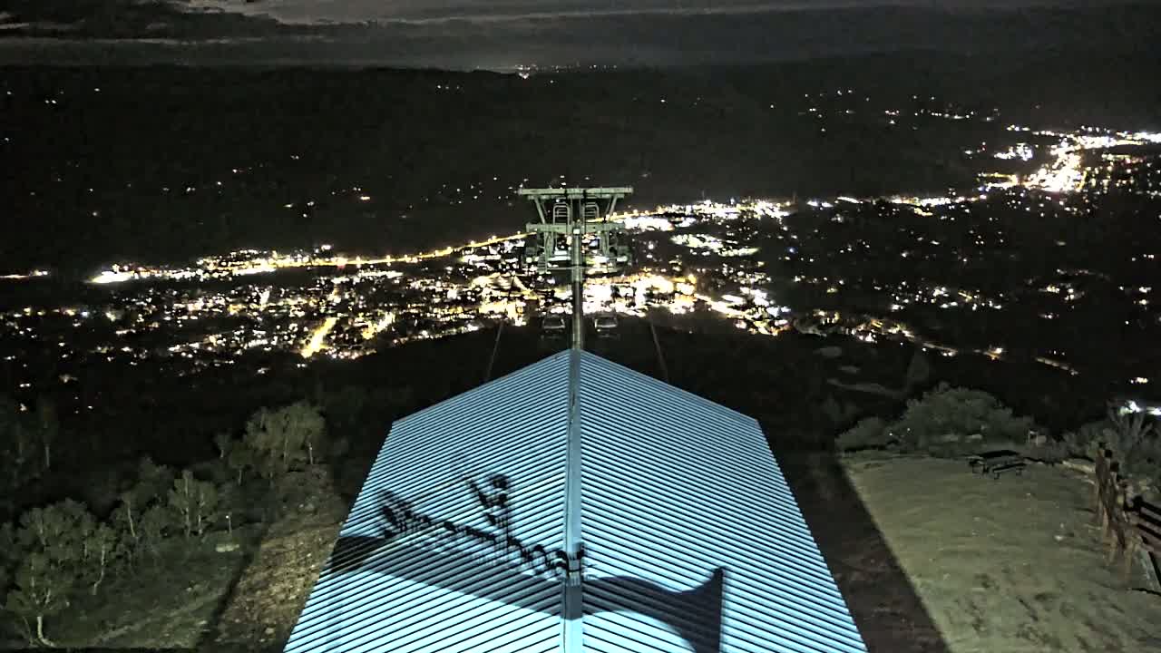A nighttime view from a hilltop shows a city illuminated below, with a chairlift and a ridged roof in the foreground.