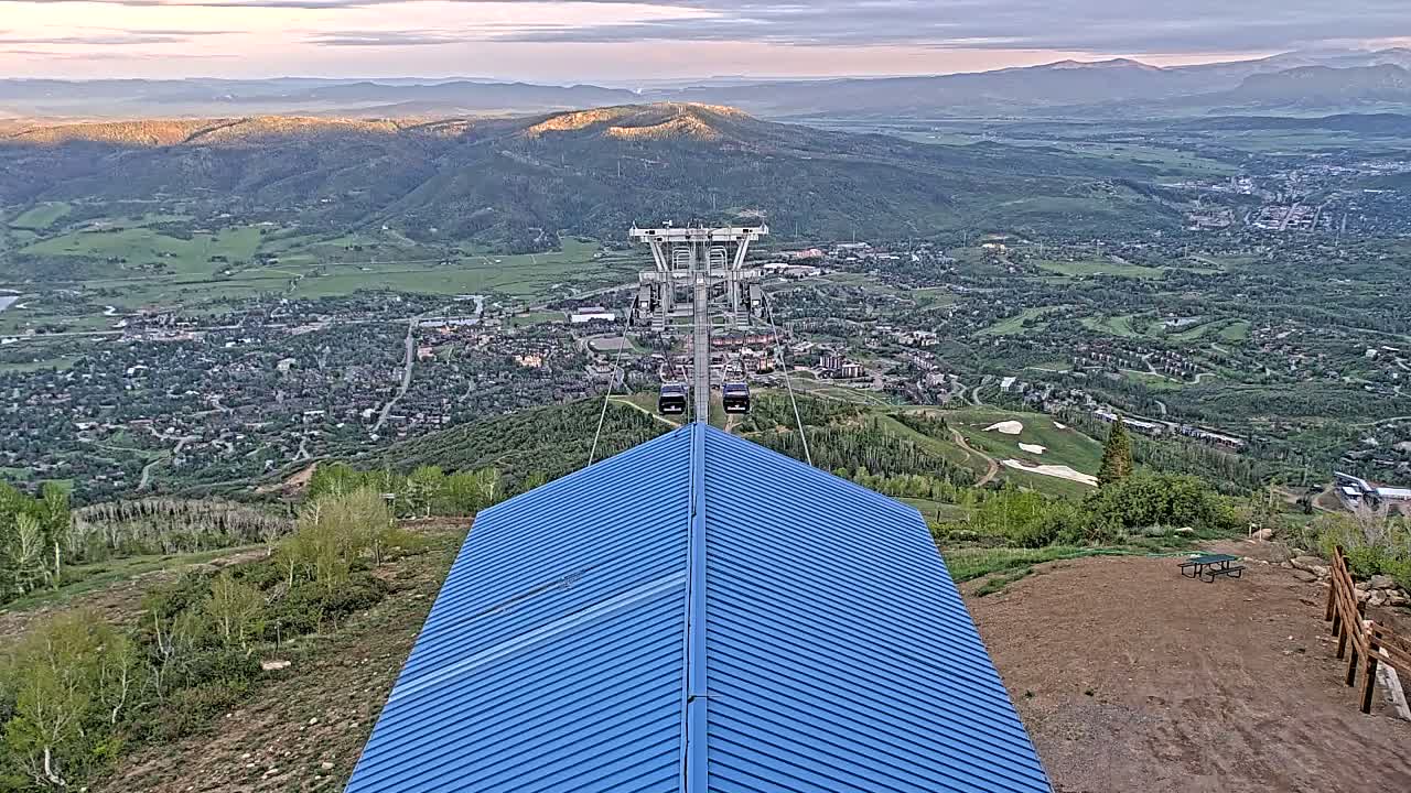 A blue-roofed building overlooks a valley town with a chairlift ascending a mountain under a partly cloudy sky.