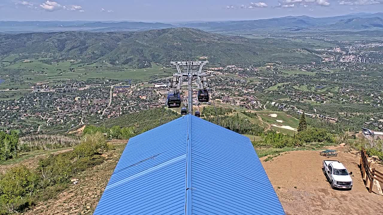 A blue-roofed building sits atop a mountain overlooking a valley town on a sunny day, with a chairlift ascending from the town in the background.
