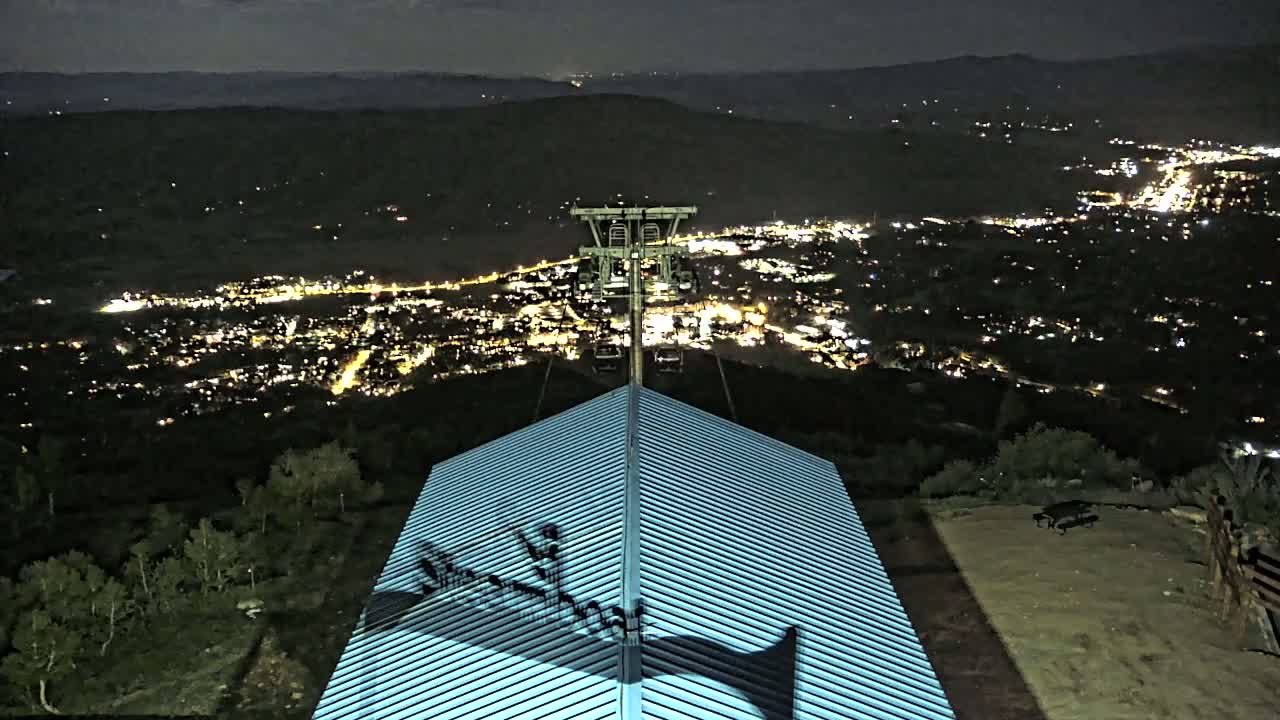 A nighttime view from atop a building with a metal roof shows a ski lift and a city illuminated below under a dark sky.